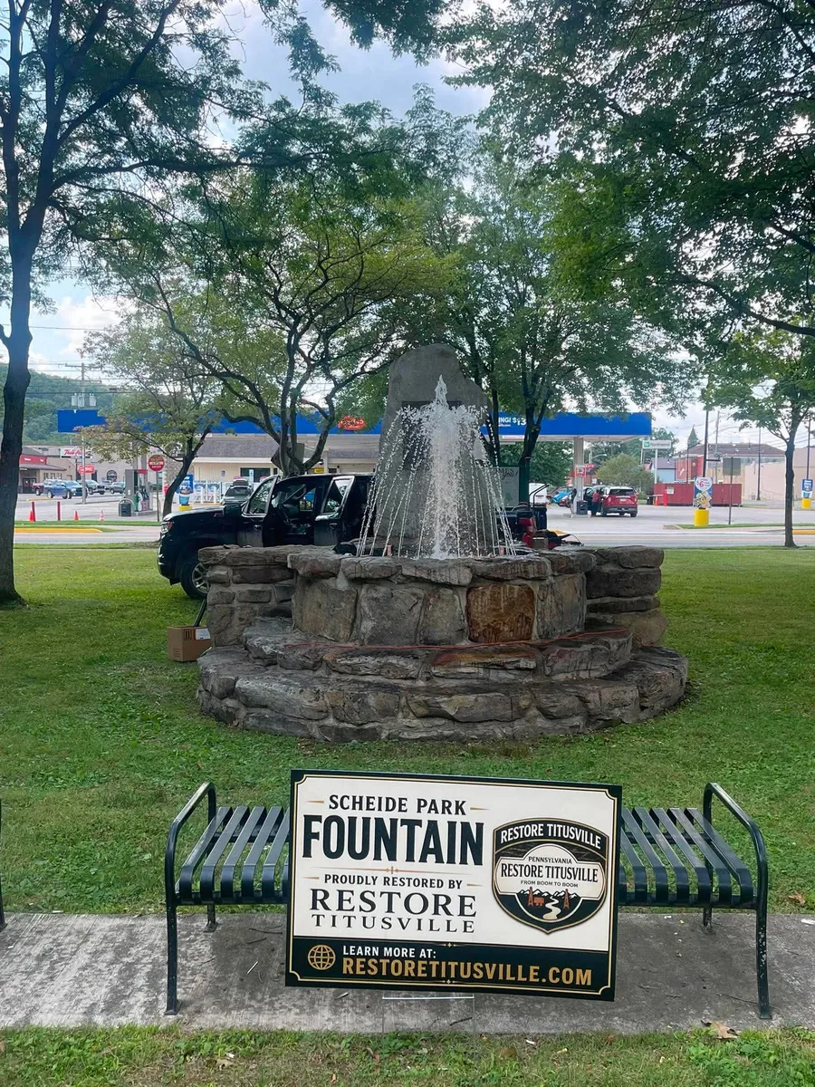 The restored stone fountain at Scheide Park running, with a Restore Titusville sign in the foreground reading "Scheide Park Fountain, proudly restored by Restore Titusville".